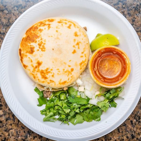 A Mexican gordita with meat, cheese, cilantro, onions, lime, and red sauce on a white plate.