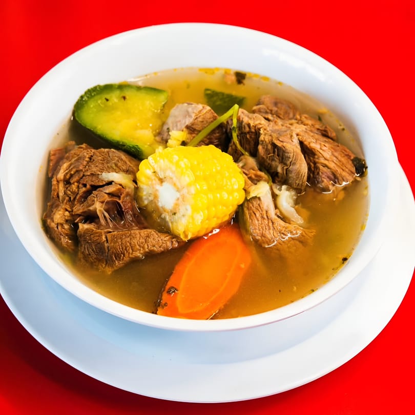 Hearty Mexican beef soup with corn, avocado, and carrots in a white bowl on a red background.