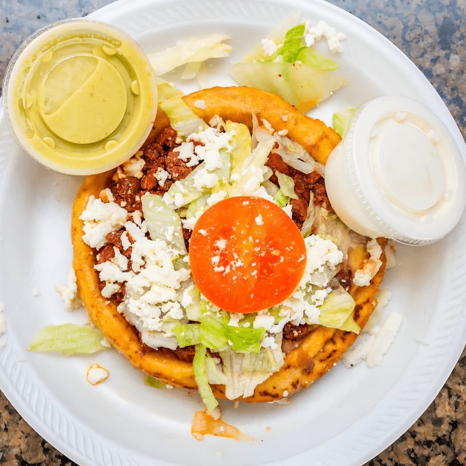 A Mexican sopes with meat, lettuce, cheese, and tomato, served with two sauce containers.
