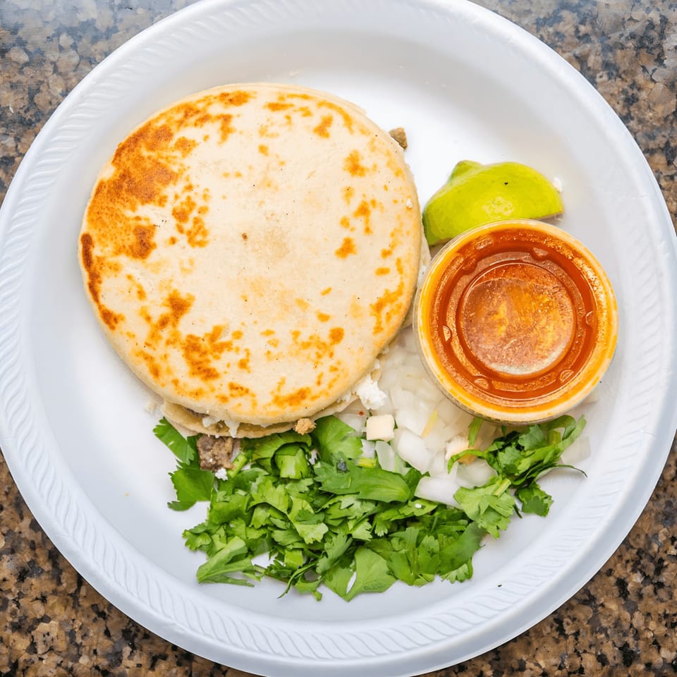 A Mexican gordita with meat, cheese, cilantro, onions, lime, and red sauce on a white plate.