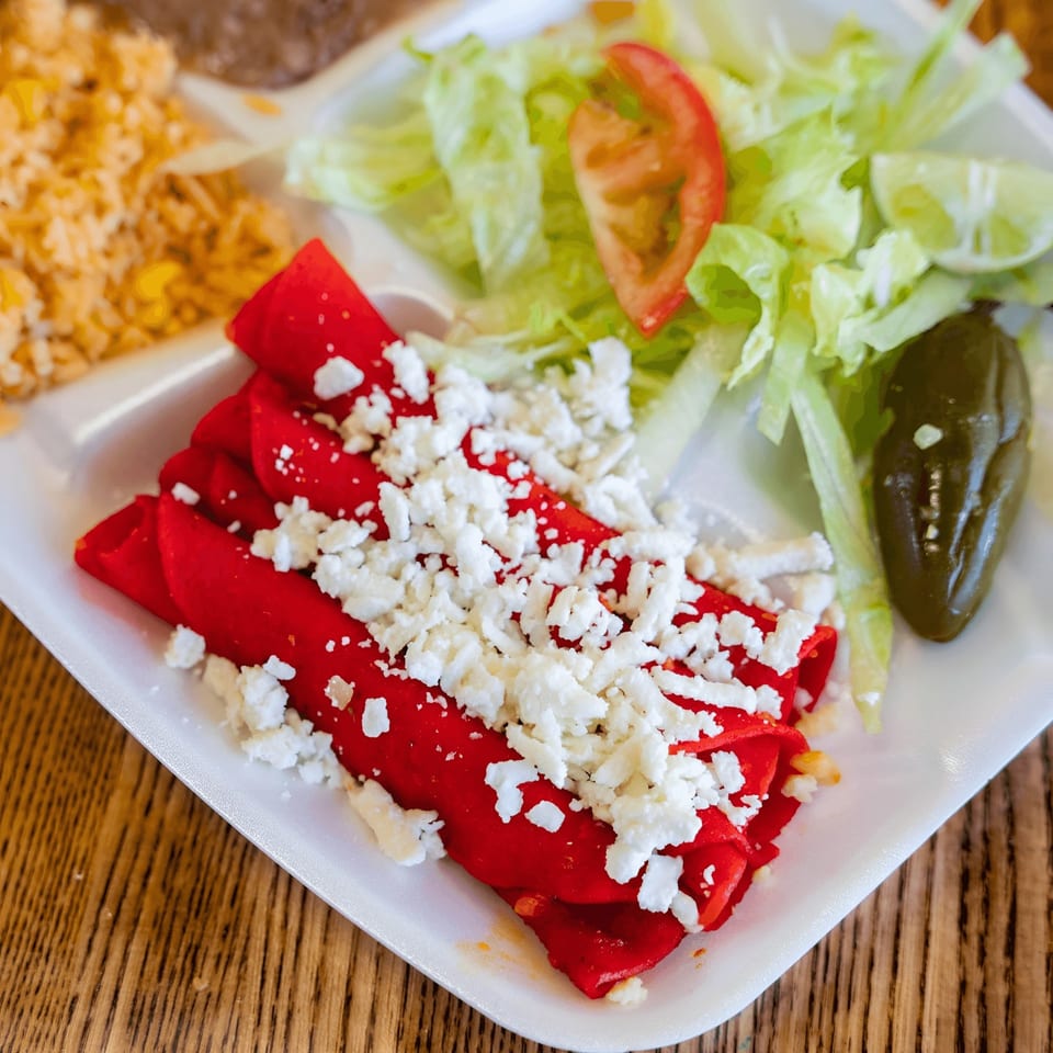 Red enchiladas topped with crumbled white cheese, served with rice, beans, lettuce, tomato, and a jalapeño.