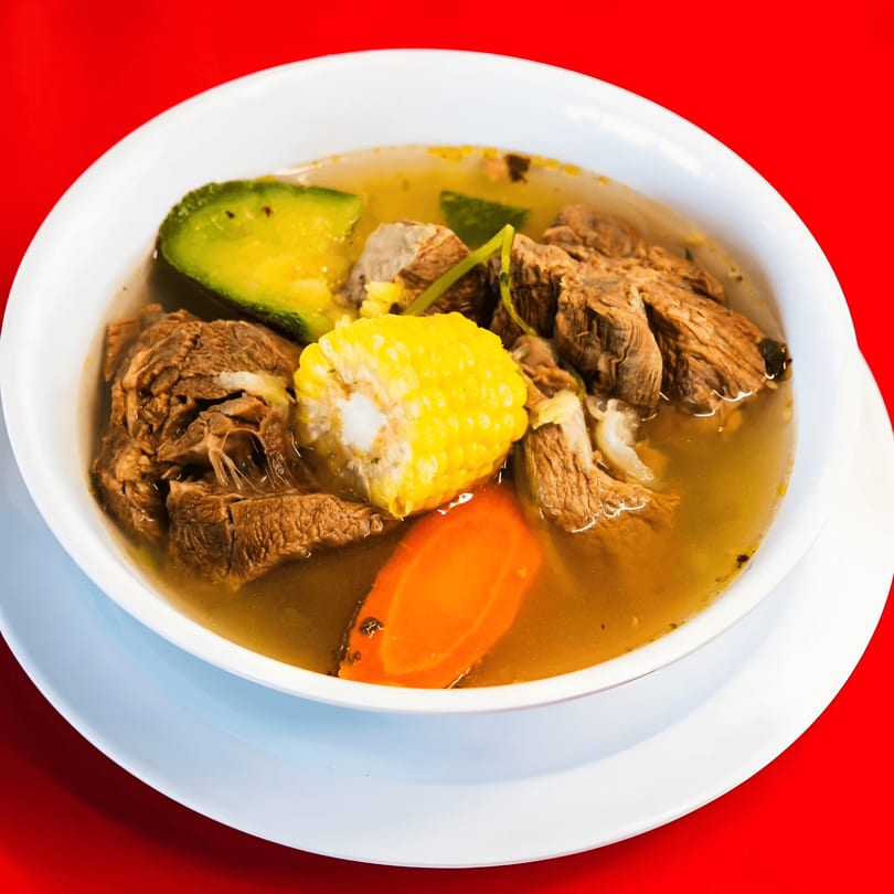 Hearty Mexican beef soup with corn, carrots, and avocado in a white bowl on a red background.